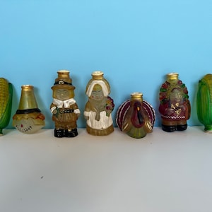 May include: Set of seven glass figurines with a Thanksgiving theme. The figurines include a pilgrim man, a pilgrim woman, a scarecrow, a turkey, and two ears of corn. The figurines are all in shades of brown, green, and red.