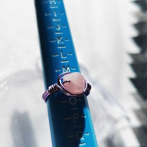 Peut inclure: Une bague en fil de fer violet avec une pierre de quartz rose. La bague est présentée sur un ajusteur de taille de bague bleu.