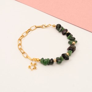 Op de afbeelding: Goudkleurige armband met een schakelketting en een gedeelte van groene en zwarte edelsteen kralen. Een kleine bedel met heldere stenen hangt aan de ketting. De armband wordt gepresenteerd tegen een witte en roze achtergrond.
