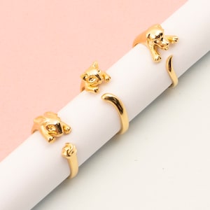 May include: Gold-coloured cat-shaped rings. The rings feature a detailed cat design, with the cat's body wrapping around the finger. The rings are displayed on a white cylindrical object against a pink and white background. The rings are a unique and stylish accessory.