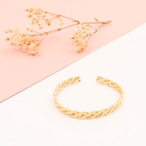 May include: A gold-coloured cuff bracelet with a braided design. The open-ended bracelet is displayed on a white and pink background, with dried flowers in the upper left corner. The bracelet is a simple, elegant piece of jewellery.