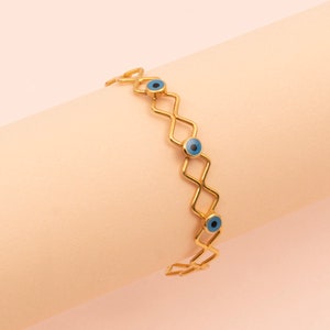 May include: Gold-coloured bracelet with a geometric design. The bracelet features diamond-shaped links, each adorned with a blue eye-shaped charm. The bracelet is displayed on a light pink surface.