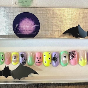 Può includere: Un set di 12 unghie finte a tema Halloween con vari design, tra cui fantasmi, zucche, ragni e occhi. Le unghie sono disposte in fila su una superficie bianca. Le unghie sono di vari colori, tra cui rosa, giallo, verde e viola.