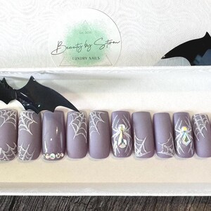 Può includere: Un set di 10 unghie finte con un design a ragnatela e un ragno su due delle unghie. Le unghie sono di colore viola chiaro e hanno una finitura opaca.