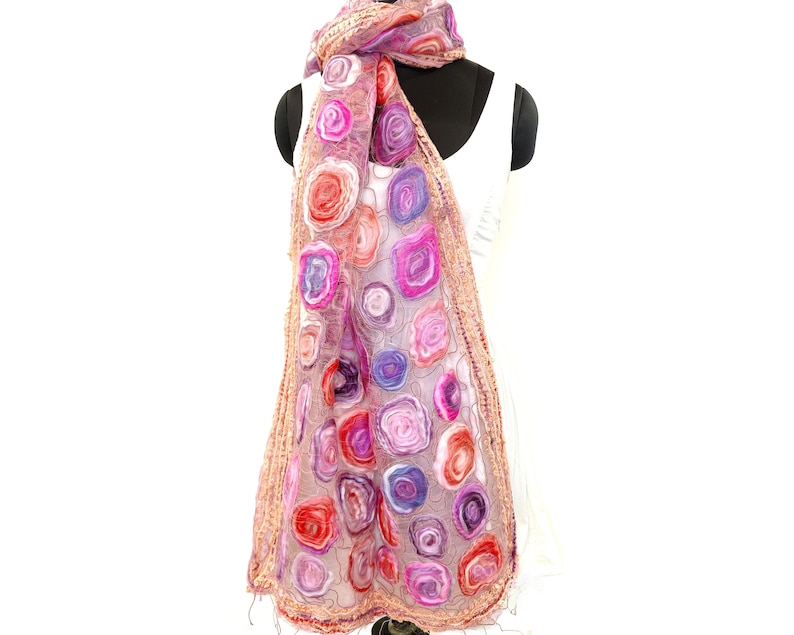 May include: A sheer, floral-patterned scarf in shades of pink, red, and purple. The scarf features a repeating pattern of stylized roses and a delicate, fringed edge. The scarf is draped over a white dress form.