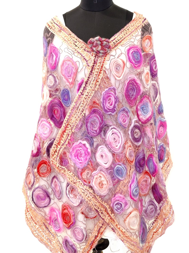 May include: A sheer, white shawl adorned with numerous felted rose designs in shades of pink, purple, and red. The shawl features a decorative border and is secured with a matching flower pin at the neckline. The shawl is draped over a mannequin.