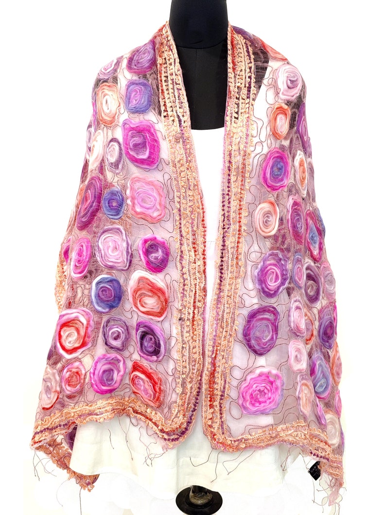 May include: A sheer, open-front shawl with a floral design. The shawl features a variety of colorful, spiral-shaped flowers in shades of pink, purple, and red. The edges are trimmed with a textured, light brown border.