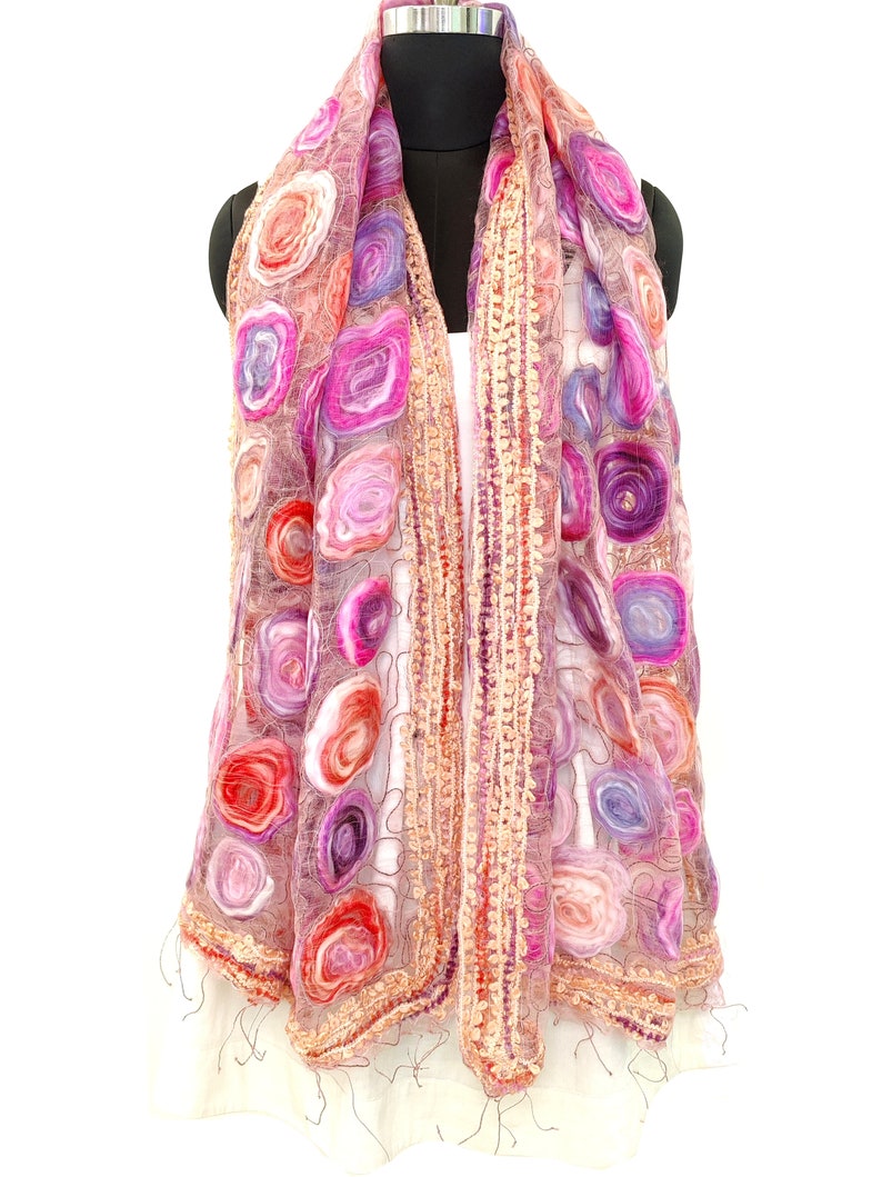 May include: A sheer, pink scarf with a floral pattern in shades of red, pink, and purple. The scarf has a delicate, open weave and is trimmed with a textured border and fringe.