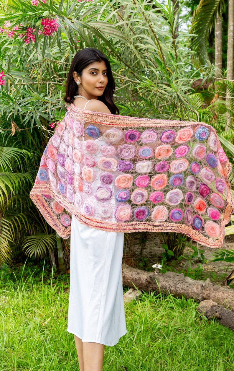 May include: A sheer, floral-patterned shawl in shades of pink, purple, and orange. The shawl is adorned with numerous fabric roses and has a delicate, crocheted border. The woman is wearing a white dress.