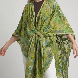 May include: A green, patterned kimono-style wrap with a tie closure. The wrap features a botanical design in shades of green, blue, and yellow. The garment is worn over a brown top and white pants.