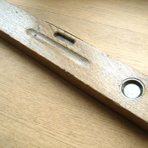 May include: A vintage wooden spirit level. The level is rectangular and features a bubble vial and a circular vial. The wood is light brown with visible grain and signs of wear, typical of an older tool.
