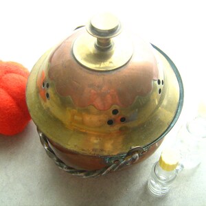 Puede incluir: Una olla vintage de cobre y latón con tapa y asa de alambre retorcido. La olla tiene un diseño decorativo alrededor del borde y pequeños agujeros en la tapa.
