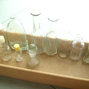 Może przedstawiać: Kolekcja dziewięciu przezroczystych szklanych butelek o różnych kształtach i rozmiarach, ułożonych w drewnianym pudełku. Butelki są puste i nie mają etykiet.