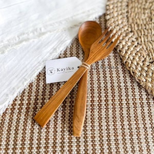 Pode incluir: Um conjunto de dois utensílios de madeira, uma colher e um garfo, feitos de madeira natural. Os utensílios estão sobre um tapete tecido marrom e branco com uma etiqueta branca que diz "Kayika Crafting the Serenity".
