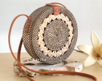 May include: A round woven crossbody bag featuring a dark brown and cream geometric pattern with a central braided cream border. The bag includes a tan leather strap and a tan leather closure tab with a small metal accent. The text Kayika is visible.