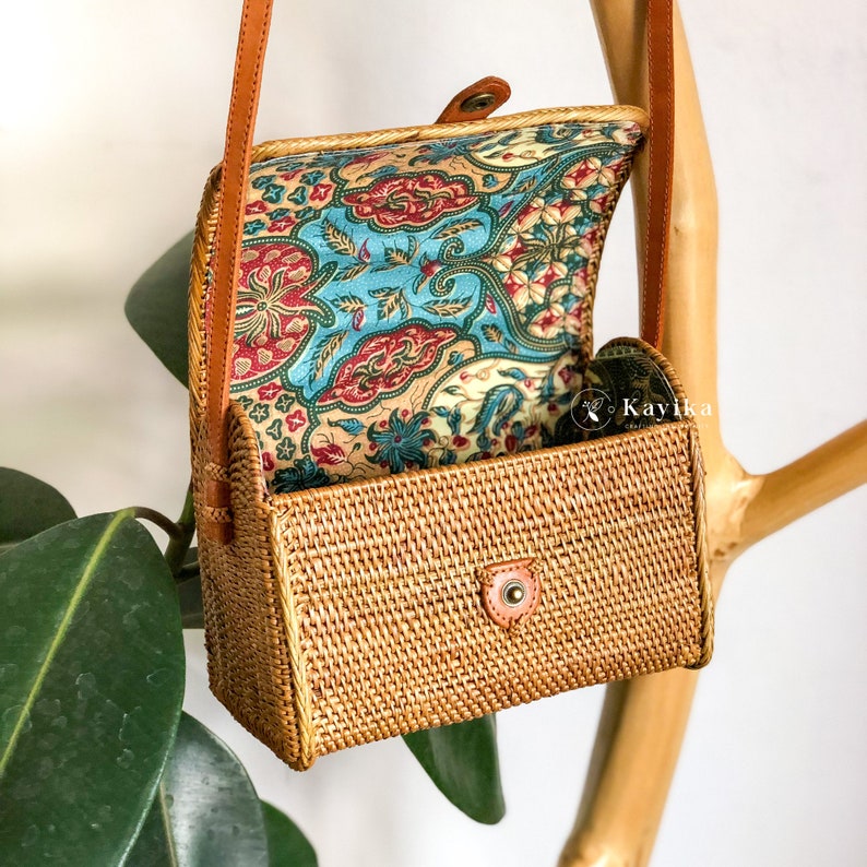 May include: A small, rectangular rattan bag with a brown leather strap and a floral patterned fabric lining. The bag is open and the lining is visible.
