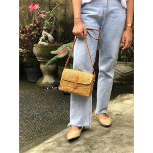 May include: A light brown woven crossbody bag with a brown leather strap and a gold buckle. The bag is worn by a person wearing light blue jeans.