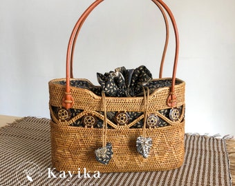 Op de afbeelding: Een geweven rotan tas met bruine leren handvatten en een zwarte en witte geprinte voering van stof. De tas heeft decoratieve houten accenten en een trekkoord sluiting.