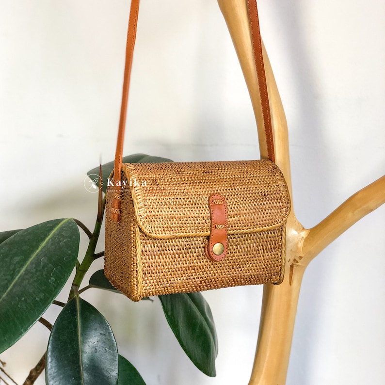 May include: A small, rectangular, woven straw bag with a brown leather strap and a brass buckle. The bag is hanging from a wooden branch.