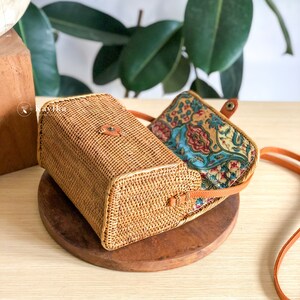 May include: A woven rattan crossbody bag with a brown leather strap and a floral patterned fabric lining.