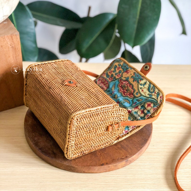 May include: A woven rattan crossbody bag with a brown leather strap and a floral patterned fabric lining.