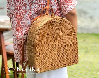 Könnte beinhalten: Ein brauner geflochtener Rattan-Rucksack mit abgerundetem Oberteil und einem Lederriemen. Der Rucksack hat ein kleines Schleifendetail oben.