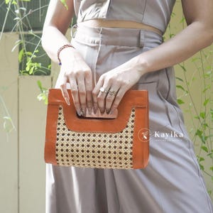Könnte beinhalten: Eine geflochtene Rattan-Clutch mit braunem Lederrahmen. Die Tasche hat einen oberen Griff und ein geflochtenes Muster.