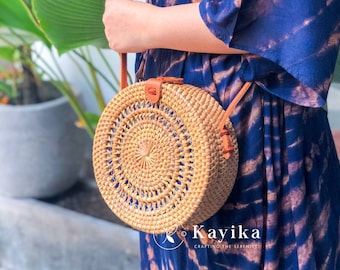 May include: A round, woven straw bag with a brown leather strap and blue accents. The bag is being held by a person wearing a blue and white tie-dye dress.