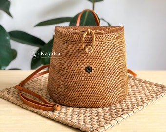 May include: A brown woven rattan backpack with a leather strap and a black cross design. The bag is sitting on a woven mat.