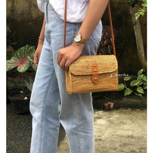 May include: A woven straw crossbody bag with a brown leather strap and flap. The bag has a brown leather buckle and a small metal accent.