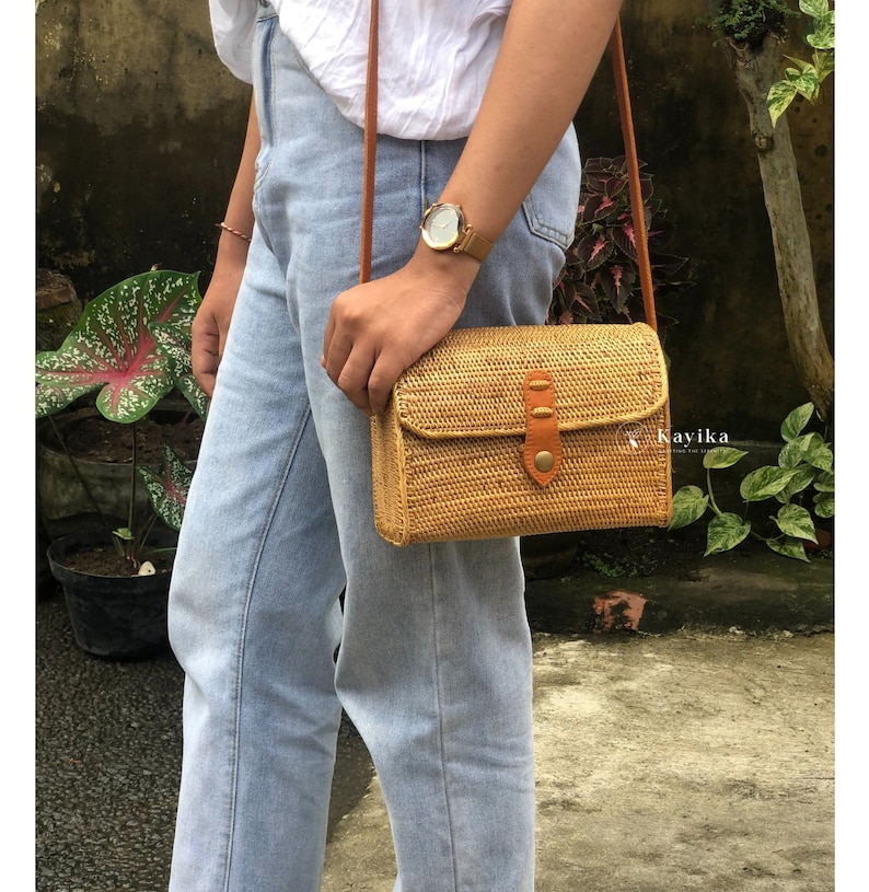 May include: A woven straw crossbody bag with a brown leather strap and flap. The bag has a brown leather buckle and a small metal accent.