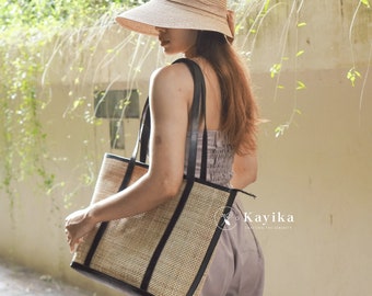 May include: A woman wearing a straw hat and a grey dress carries a woven tote bag with black leather trim. The bag has a woven pattern and black leather straps.