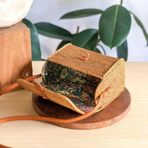 May include: A small, rectangular, woven rattan bag with a brown leather strap. The bag is open, revealing a floral patterned fabric lining.