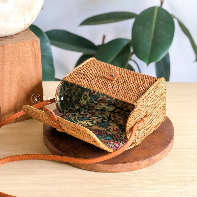 May include: A small, rectangular, woven rattan bag with a brown leather strap. The bag is open, revealing a floral patterned fabric lining.