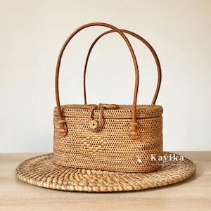 Könnte beinhalten: Eine rechteckige, gewebte Rattan-Handtasche mit zwei gewölbten braunen Ledergriffen. Die Tasche hat ein gewebtes Muster und einen Knopfverschluss. Sie steht auf einem runden, gewebten Platzset. Der Markenname "Kayika" ist sichtbar.