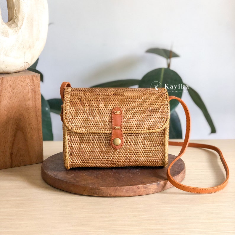 May include: A small, rectangular woven bag with a brown leather strap and a brass buckle. The bag is made of natural materials and has a light brown color.