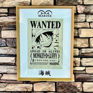 Op de afbeelding: Een gezochtposter voor Monkey D. Luffy uit de anime One Piece. De poster is ingelijst en toont een cartoonbeeld van Luffy met de tekst "WANTED" en "DEAD OR ALIVE?"  De poster bevat ook de tekst "MONKEY-D.LUFFY" en een beloning van "$1.500.000.000-." De poster hangt aan een bakstenen muur.
