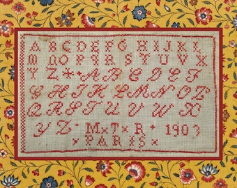 Op de afbeelding: Een vintage kruissteek sampler met het alfabet in rode draad op een lichtgrijze stof. De sampler is omlijst met een rode rand en geplaatst tegen een gele bloemenachtergrond. De tekst bevat het jaartal 1903 en het woord PARIS.