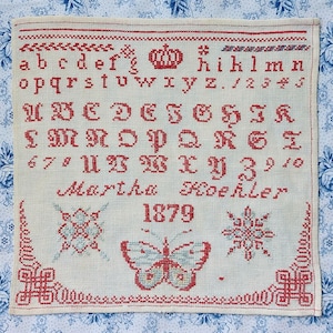 May include: A vintage cross-stitch sampler with red and blue embroidery on a cream-colored fabric. The sampler features the alphabet, numbers, a crown, a butterfly, and the name "Martha Koehler" and the year "1879".