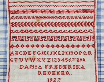 May include: A white linen sampler with red embroidery. The sampler includes the alphabet, numbers, and the name "DANIA FREDERIKA REDEKER." The date "1927" is also embroidered on the sampler.