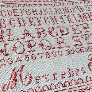 May include: A white fabric sampler with red cross-stitch embroidery. The sampler features the alphabet, numbers, and the word "Getredes".