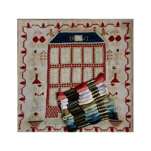 May include: A vintage needlepoint sampler featuring a red brick house with a blue roof and a white door. The house has 1861 written above the door. The sampler is surrounded by a red and white border with a floral design. A stack of embroidery floss in various colors is in the bottom right corner of the image.