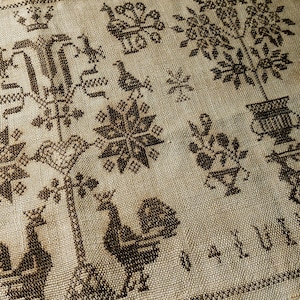 May include: A close-up of a brown and white linen fabric with a black embroidered design. The design features a variety of motifs, including birds, flowers, and geometric shapes.