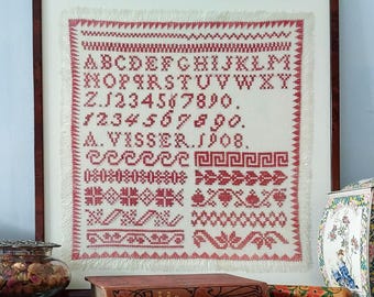 Op de afbeelding: Een ingelijst wit stoffen sampler met rode borduurwerk. De sampler bevat het alfabet, cijfers en verschillende decoratieve steken. De sampler is gesigneerd met "A. Visser. 1908."