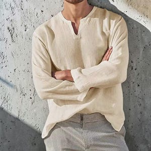 Puede incluir: Una camisa de manga larga en color crema con escote en pico y acabado de tela texturizada. La camisa presenta un corte relajado con mangas rectas y se combina con pantalones de rayas verticales grises y blancas con una cinturilla visible.