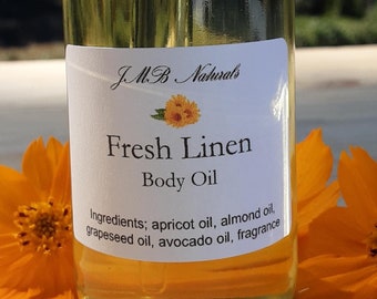 May include: A glass bottle of Fresh Linen Body Oil with a dropper top. The bottle is sitting on a wooden surface with two yellow flowers on either side. The label on the bottle reads "JMB Naturals Fresh Linen Body Oil" and lists the ingredients: apricot oil, almond oil, grapeseed oil, avocado oil, and fragrance.