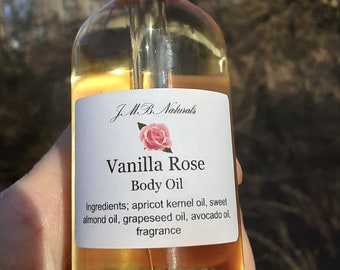 May include: A glass bottle of Vanilla Rose Body Oil with a dropper top. The label is white with black text and a pink rose graphic. The ingredients are listed as apricot kernel oil, sweet almond oil, grapeseed oil, avocado oil, and fragrance.