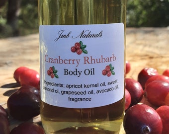 Puede incluir: Una botella de vidrio de aceite corporal de arándano y ruibarbo con una tapa negra. La botella está sobre una superficie de madera rodeada de arándanos rojos. La etiqueta dice "Jmb Naturals Cranberry Rhubarb Body Oil" y enumera los ingredientes: aceite de hueso de albaricoque, aceite de almendras dulces, aceite de semilla de uva, aceite de aguacate y fragancia.