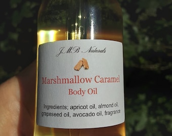 May include: A clear glass bottle with a black dropper top and a white label. The label reads "JMB Naturals Marshmallow Caramel Body Oil" and lists the ingredients: apricot oil, almond oil, grapeseed oil, avocado oil, and fragrance.