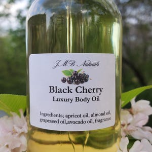 Op de afbeelding: Een heldere glazen fles Black Cherry Luxury Body Oil met een zwarte dop. Het etiket bevat de merknaam, productnaam en ingredi&euml;nten. Witte bloemen en groene bladeren omringen de fles.